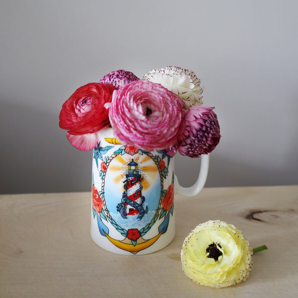 Small white bine china jug with lighthouse and kraken design inspired by sailor's tattoo's. The jug is filled with dainty pink and red ranunculus and is sitting on a wooden table top against a grey background. There is a yellow flower laying on the wooden surface just to the right of the jug.