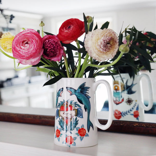 White bone china jug decorated with shark & heart tattoo inspired design. The jug is filled with red, pink & yellow ranunculus and is reflected in the mirror behind it.