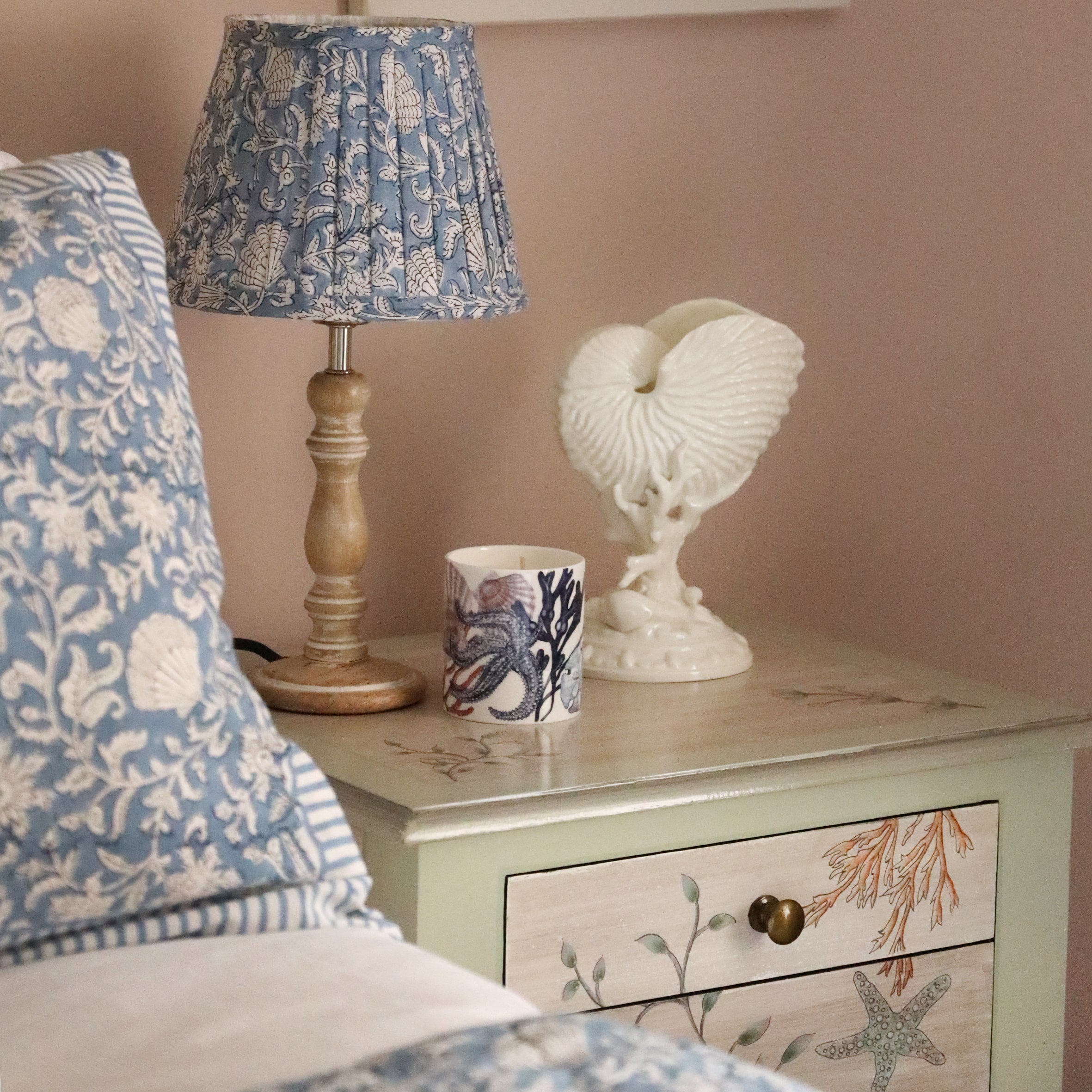 Small pleated Atlantic Blue Seashell Flower lampshade in hand blocked print in blue and white on a wooden lampbase on a bedside cabinet.On the cabinet is a beachcomber candle and a shell ornament.In the foreground you can see our hand block printed bed linen on a bed.