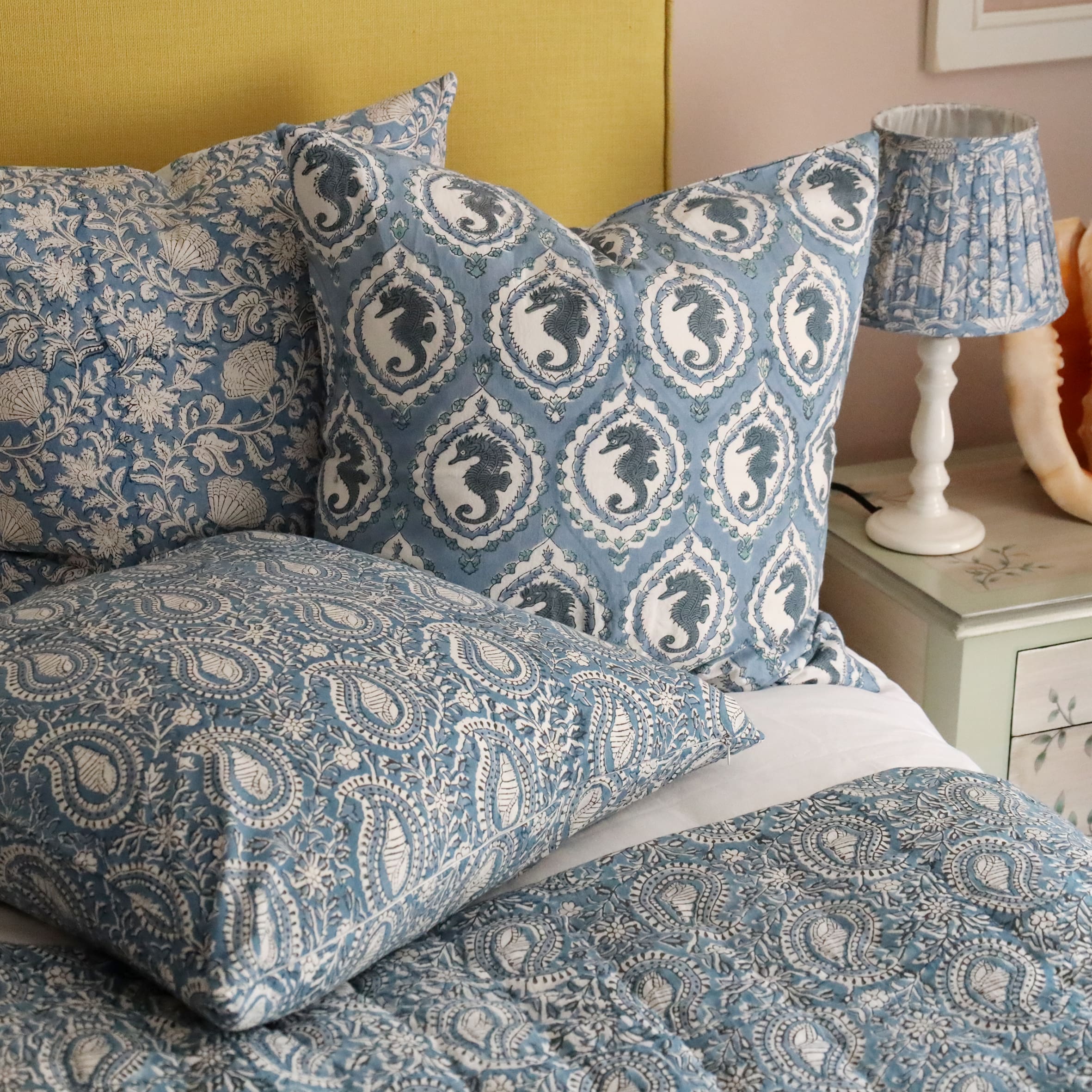 Ocean Blue Seahorse Cameo cushion which is Hand block printed fabric .Behind the cushion is another Handblock printed cushion,both on a bed.Next to the bed is a bedside cabinet beautifully decorated with sea creatures,on that is a lamp base with a handblock printed pleated lampshade. 