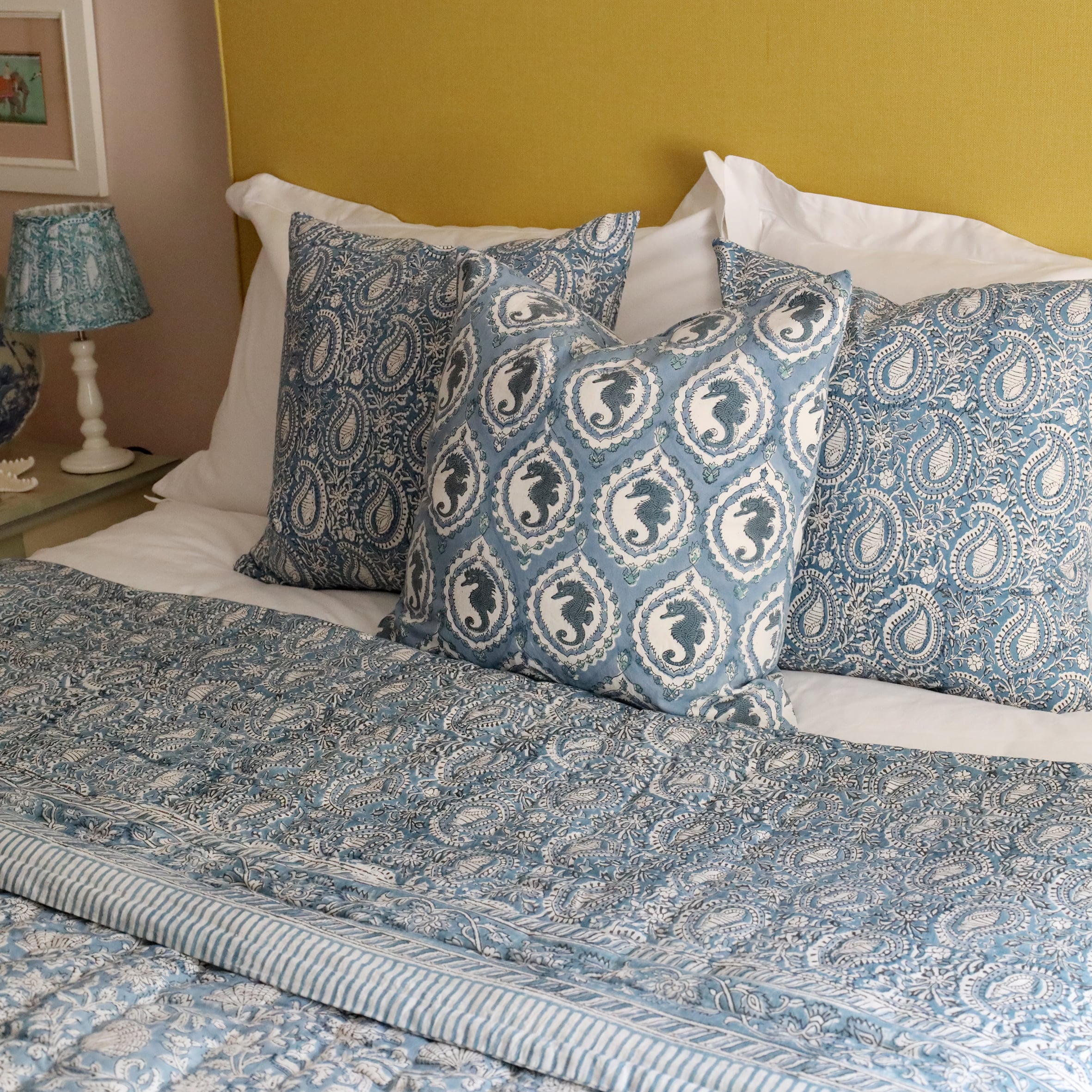 Ocean Blue Seahorse cushion which is Hand block printed fabric in a  blue .The cushion is placed on a bed with two other block print cushions behind in on a crisp white bed,in front of a mustard yellow headboard.Next to the bed is a pink lampbase with a pleated block print fabric lampshade on a hand painted bedside table