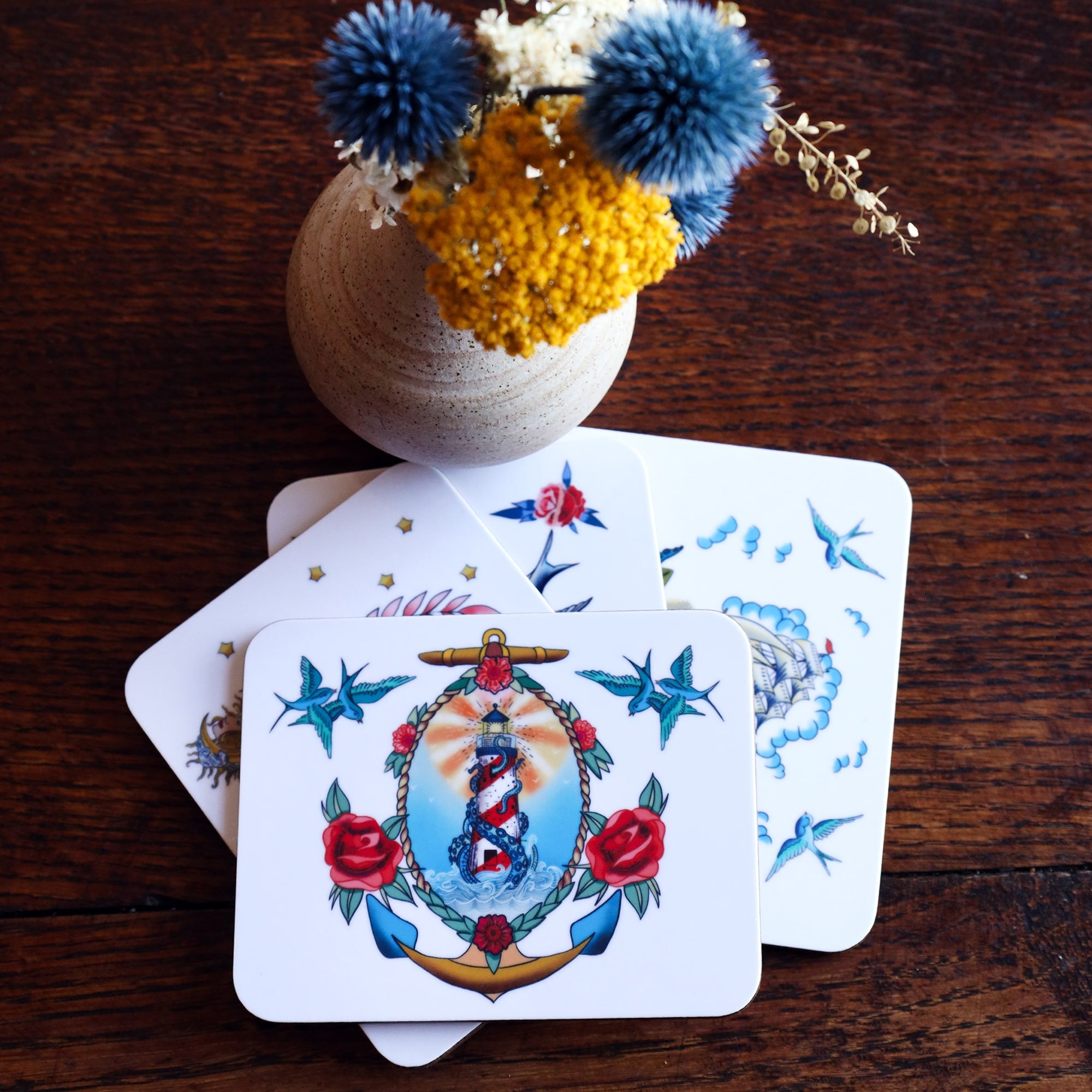 4 melamine coasters with sailors tattoo designs in a pile on dark wood table top with a small vase with dried flowers. Only the top coaster is fully visible with lighthouse and kraken design surrounded by swallows and roses.
