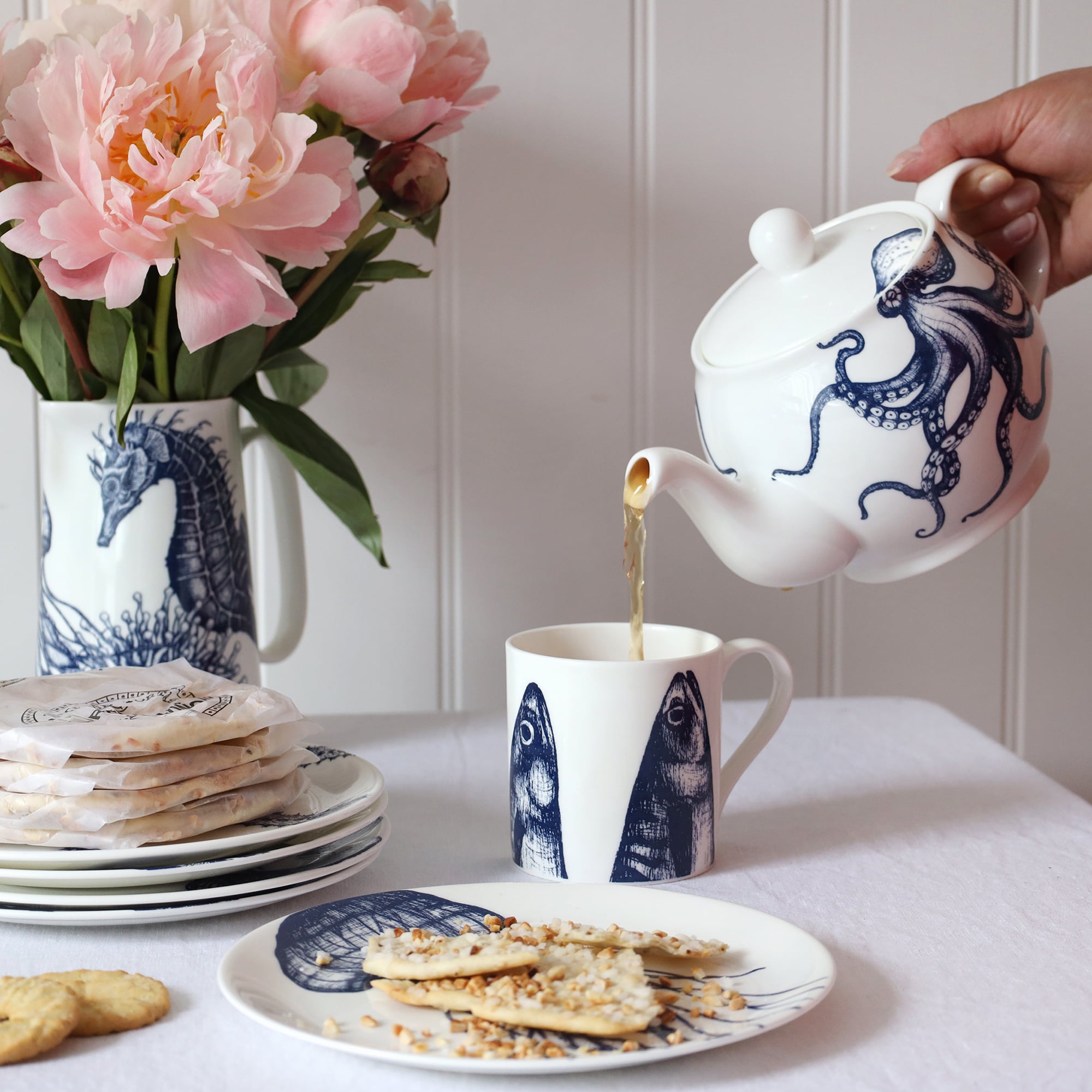 An informal table setting with blue & white coastal bone china on white linen and a shiplap background. There is an teapot with an octopus design pouring tea into a white mug that is decorated with a dark blue illustrated mackerel heads design.