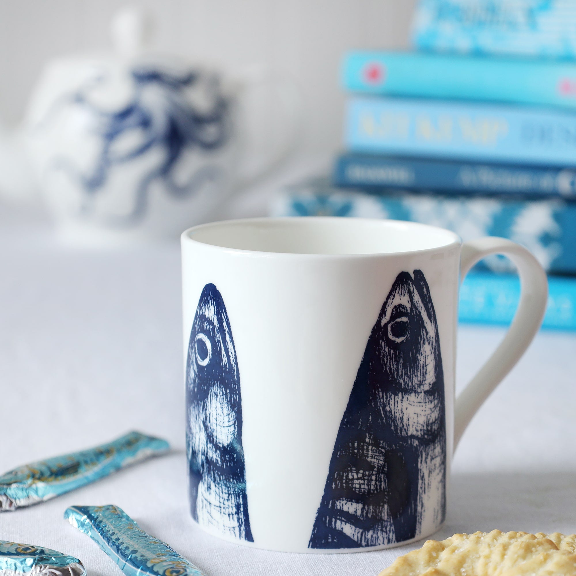 A close up of a white bone china mug with dark blue illustrated mackerel heads design coming up from the bottom of the mug. This is sitting on a white table cloth with some chocolate sardines and an octopus teapot & pile of blue books int he background.