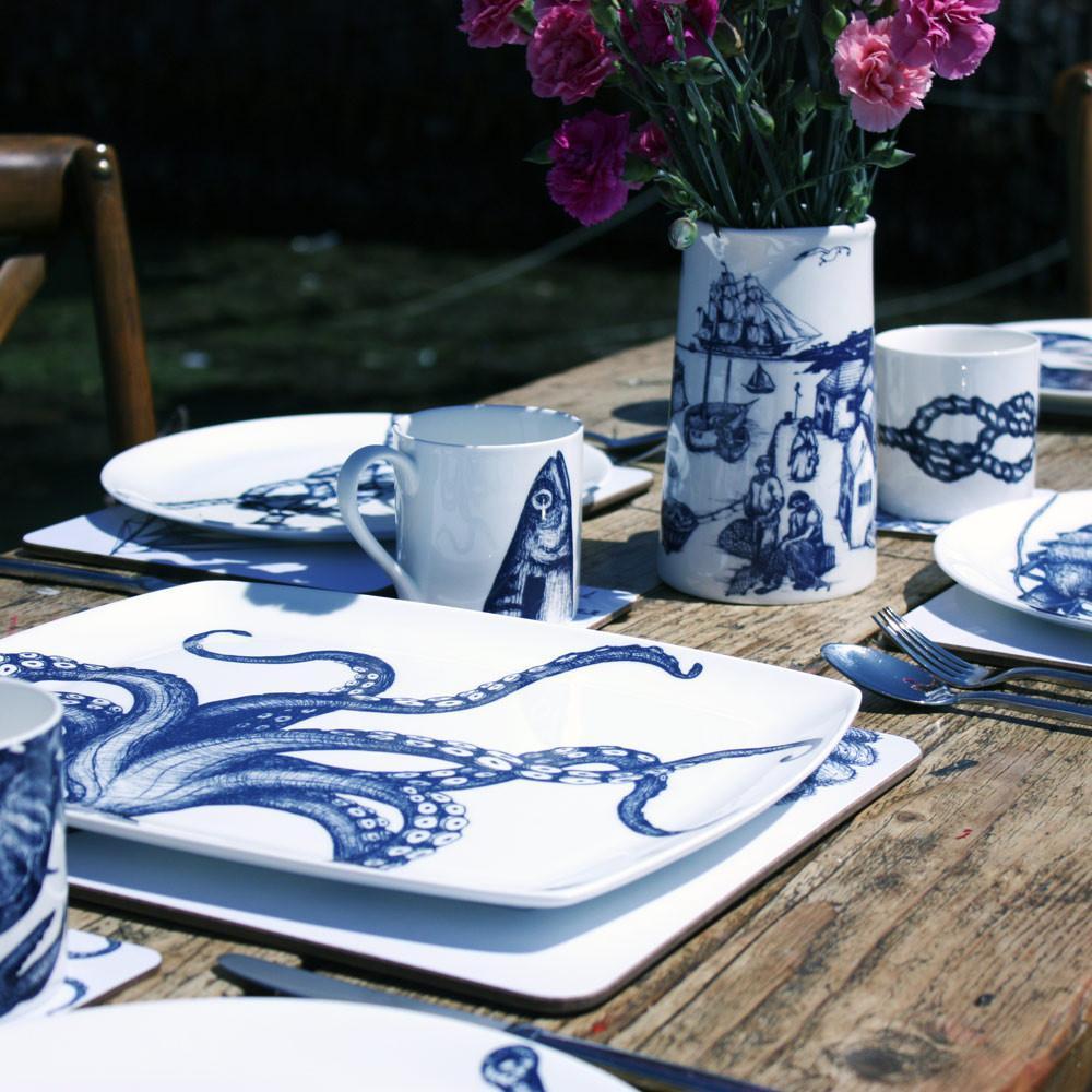 Bone china white mug featuring hand drawn Mackerel Heads design in classic navy on a wood table in an outdoor setting.On the table are other Cream Cornwall white bone china products including a large Harbour scene Jug with pink carnations ,Octopus Platter and placemats on a wooden table
