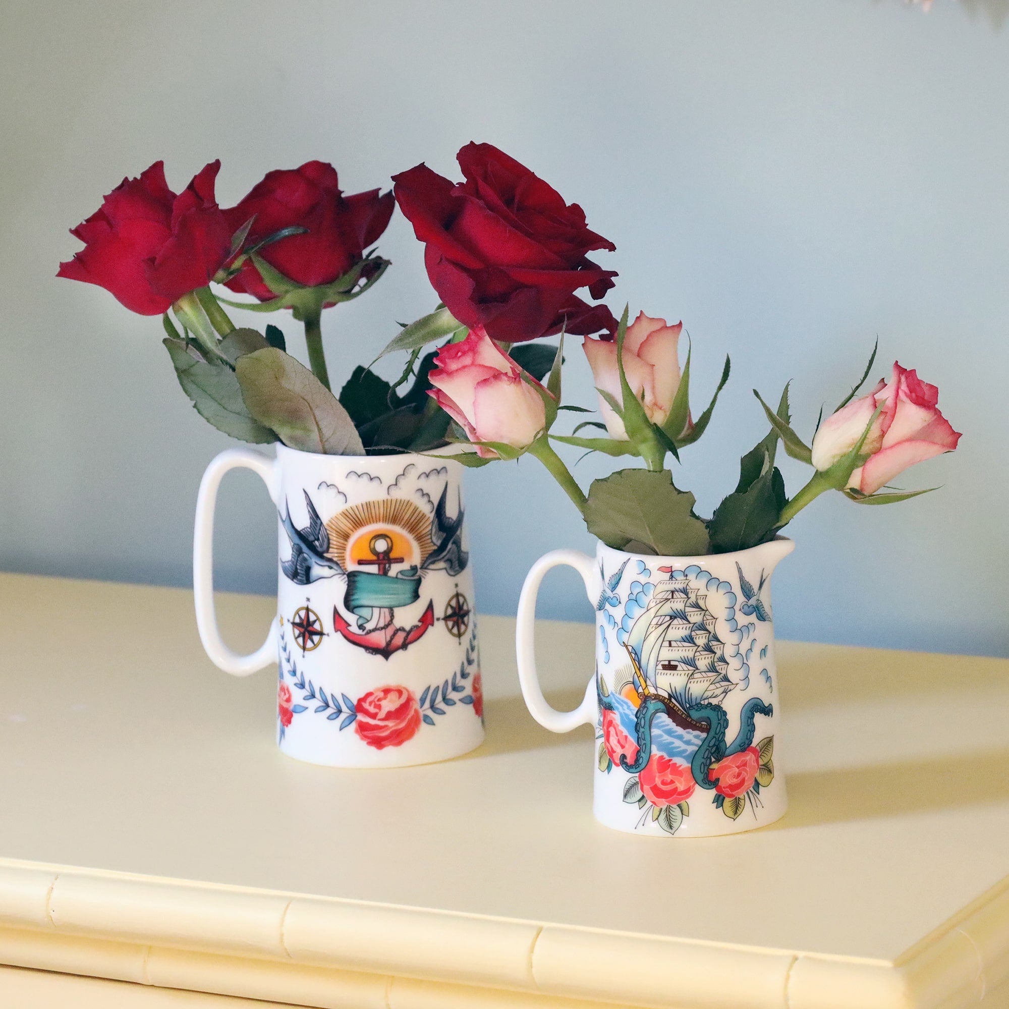 2 white jugs decorated in brightly coloured tattoo inspired patterns of swallow and anchors, ship and kraken and roses. Both are filled with roses one with red and the other with pink and white, sitting on a soft yellow set of drawers and a soft blue wall behind.