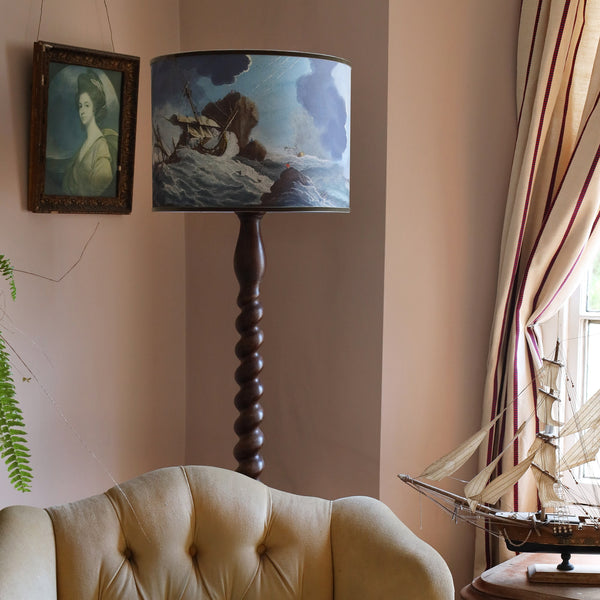 A shipwreck scene lampshade in blues and brown on a dark wooden twisted standard lamp base, placed behind a mustard velvet buttoned high back chair with an old portrait of a lady to the side and a model of a galleon on the table, all set against pink plaster walls. 