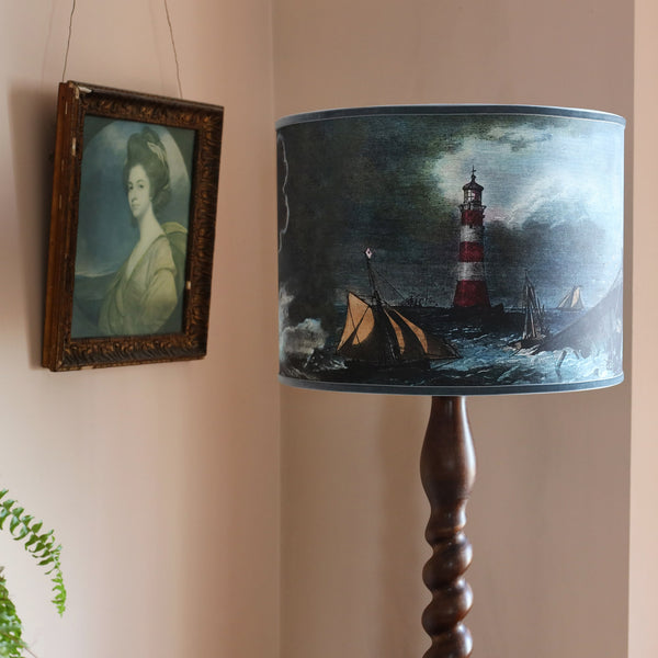 A shipwreck scene & lighthouse lampshade on a dark wooden twisted standard lamp base, placed behind a mustard velvet buttoned high back chair with an old portrait of a lady to the side and a model of a galleon on the table, all set against pink plaster walls. 