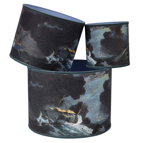 A stack of 3 different sized lampshades on white background, with nautical seascape images of a shipwreck in stormy seas in dark blues and browns and trimmed with a blue grey velvet ribbon.