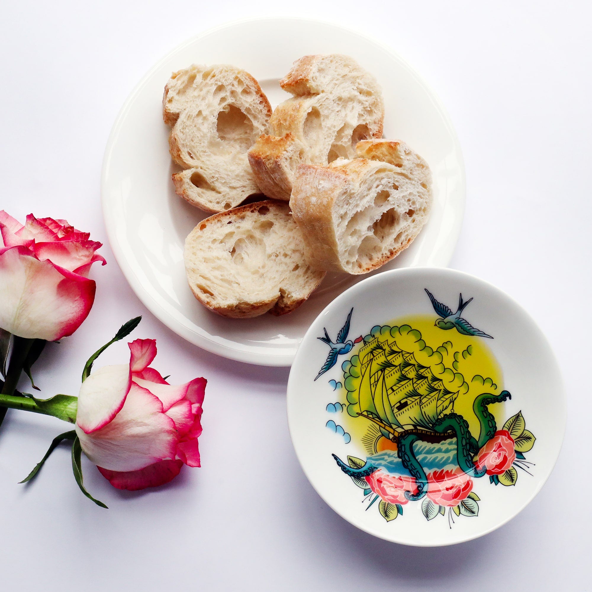 Nibbles dish with ship and kraken design in the style of a tattoo, with olive oil in and a plate of bread next to it. There are 2 pink & white roses to the side.