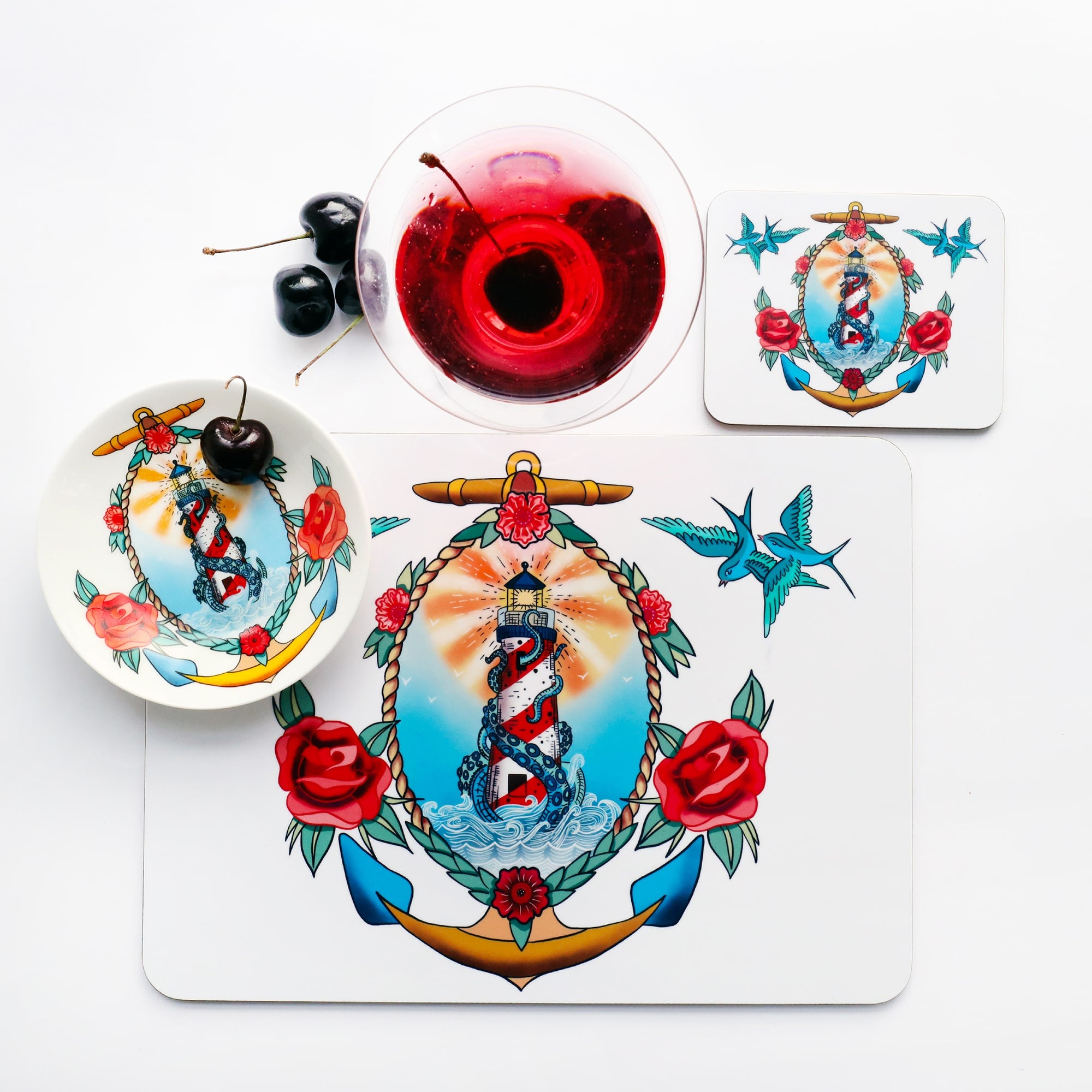 Place setting with placemat, coaster and nibbles dish all the the same lighthouse and kraken design that is inspired by a sailors tattoos. The place setting is shot from above and there is a red cocktail and cherries just above the placemat.