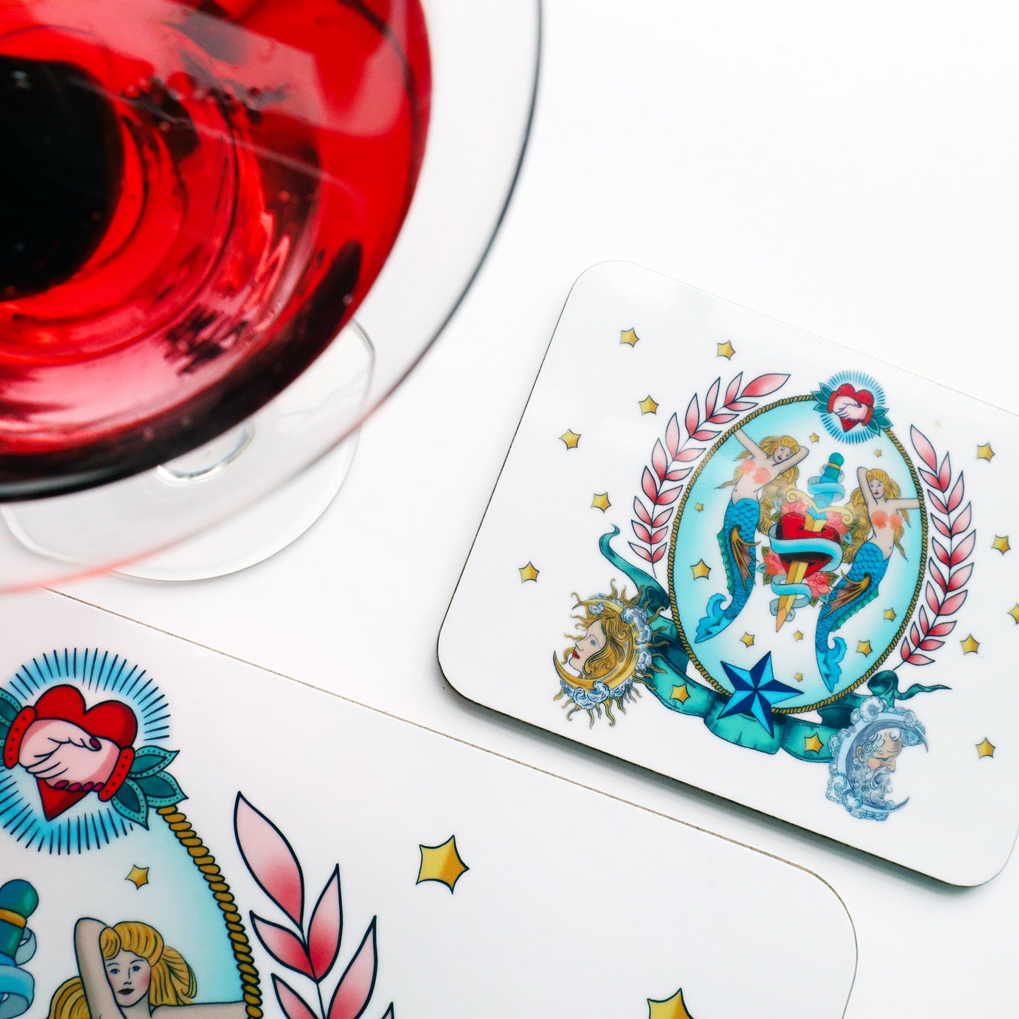 Melamine coaster with a sailors tattoos inspired illustration of mermaids, heart and dagger in a vignette of pink laurel, rope and sun and moon faces. There is the corner of the matching placemat just visible as well as a red cocktail in the top left corner. 