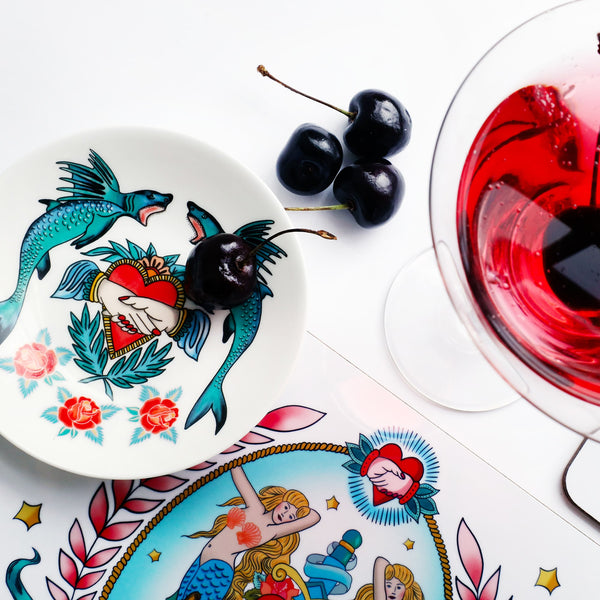 Nibbles dish decorated with sharks, heart and hands and roses in a tattoo style. The dish is on a placemat with a mermaid design and is just visible along with a cocktail glass and 3 cherries.