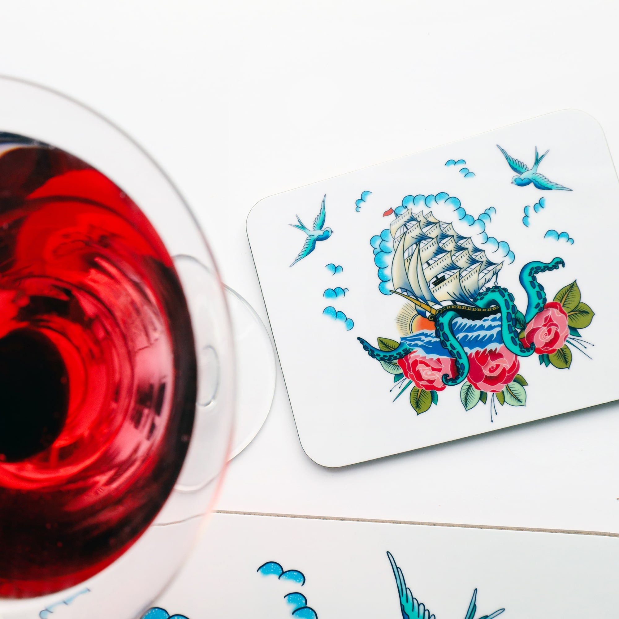 Ship and kraken design coaster inspired by sailors tattoos. This is on a white background with the top of a matching placemat just visible at the bottom and a red cocktail to the left.