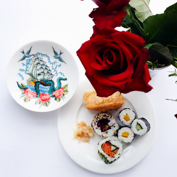 Nibbles dish with ship and kraken design in a tattoo style, sitting next to a plate of sushi and red roses.