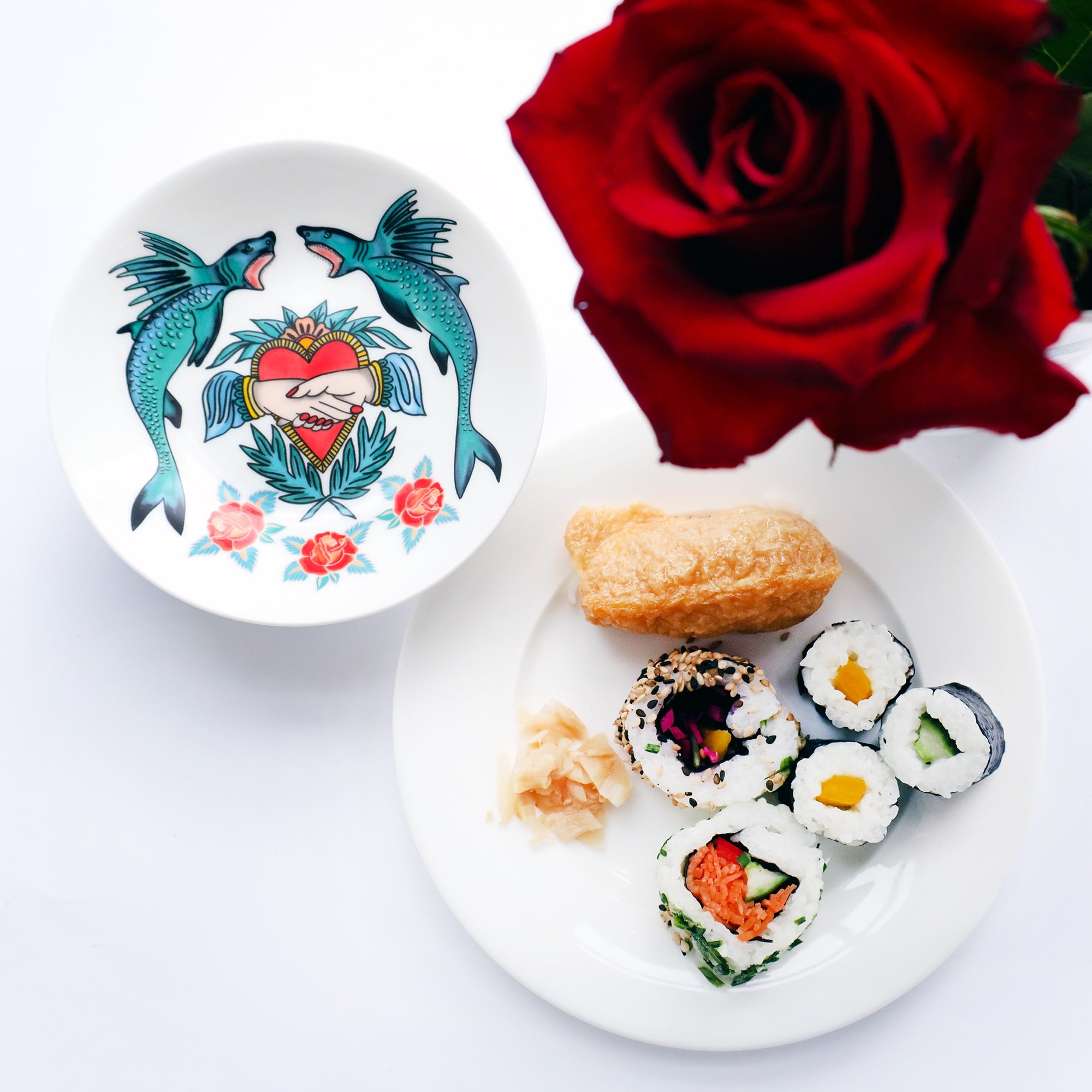 Nibbles dish with sharks, heart and hand and roses in a tattoo style. there is a plate of sushi next to it and a red rose.