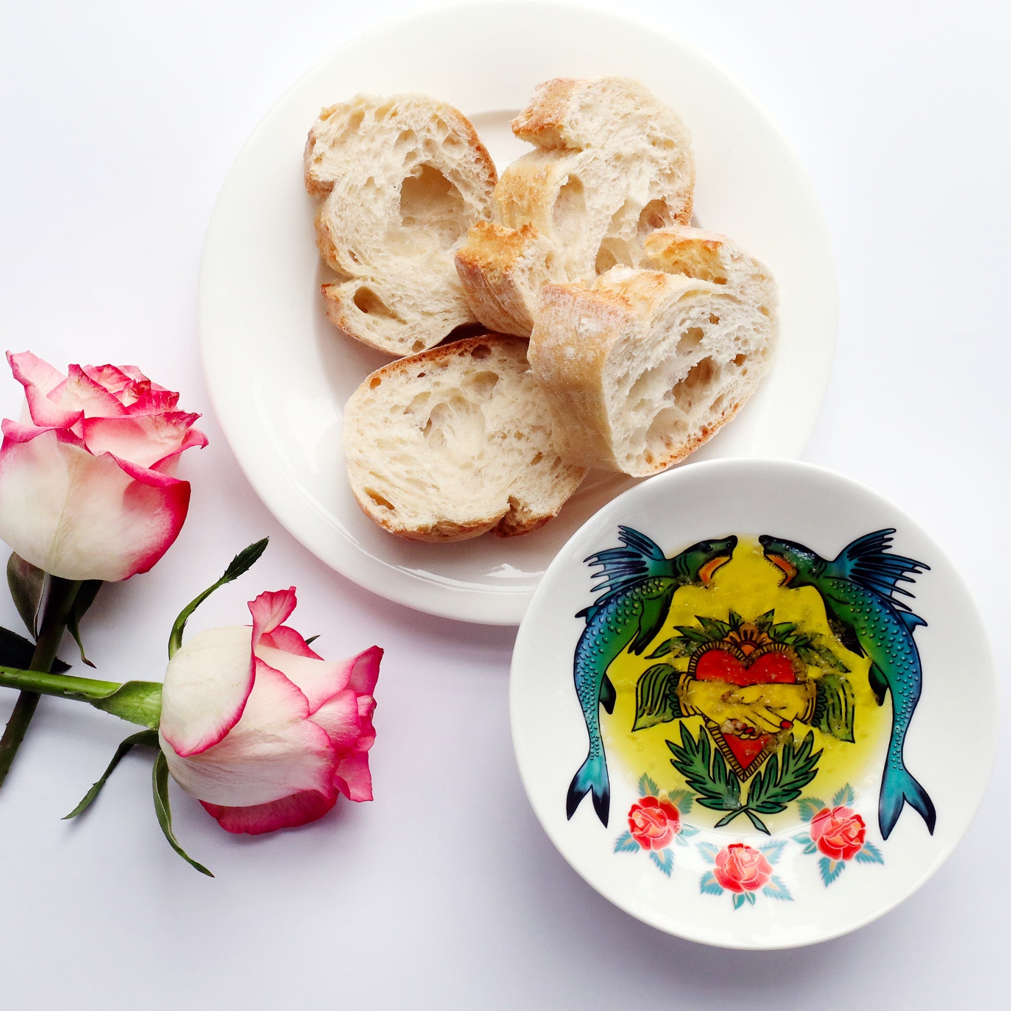 Nibbles dish decorated in shark, heart and roses design inspired by tattoos, filled with olive oil and salt. There is a plate of bread next to the dish and 2 pink and white roses.