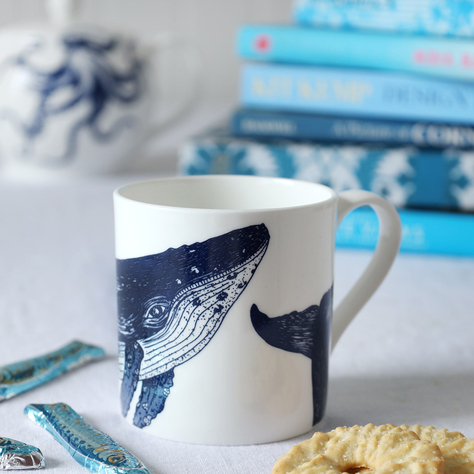 Close up of a white bone china mug with a blue illustrated humpback whale on it, sitting on a table with biscuits and sardine chocolates and a pile of out of focus blue books and teapot in the background. 