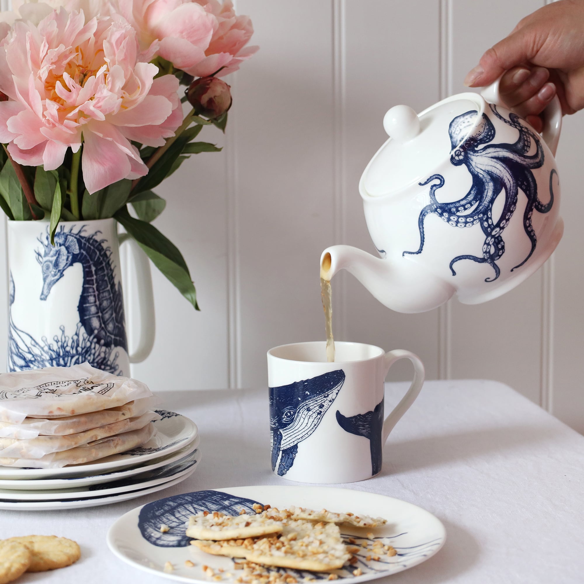 A blue & white informal; table setting set against a white shiplap wall and linen tablecloth. An octopus design adorns a teapot which is pouring tea into a white bone china mug decorated with a blue illustrated humpback whale..