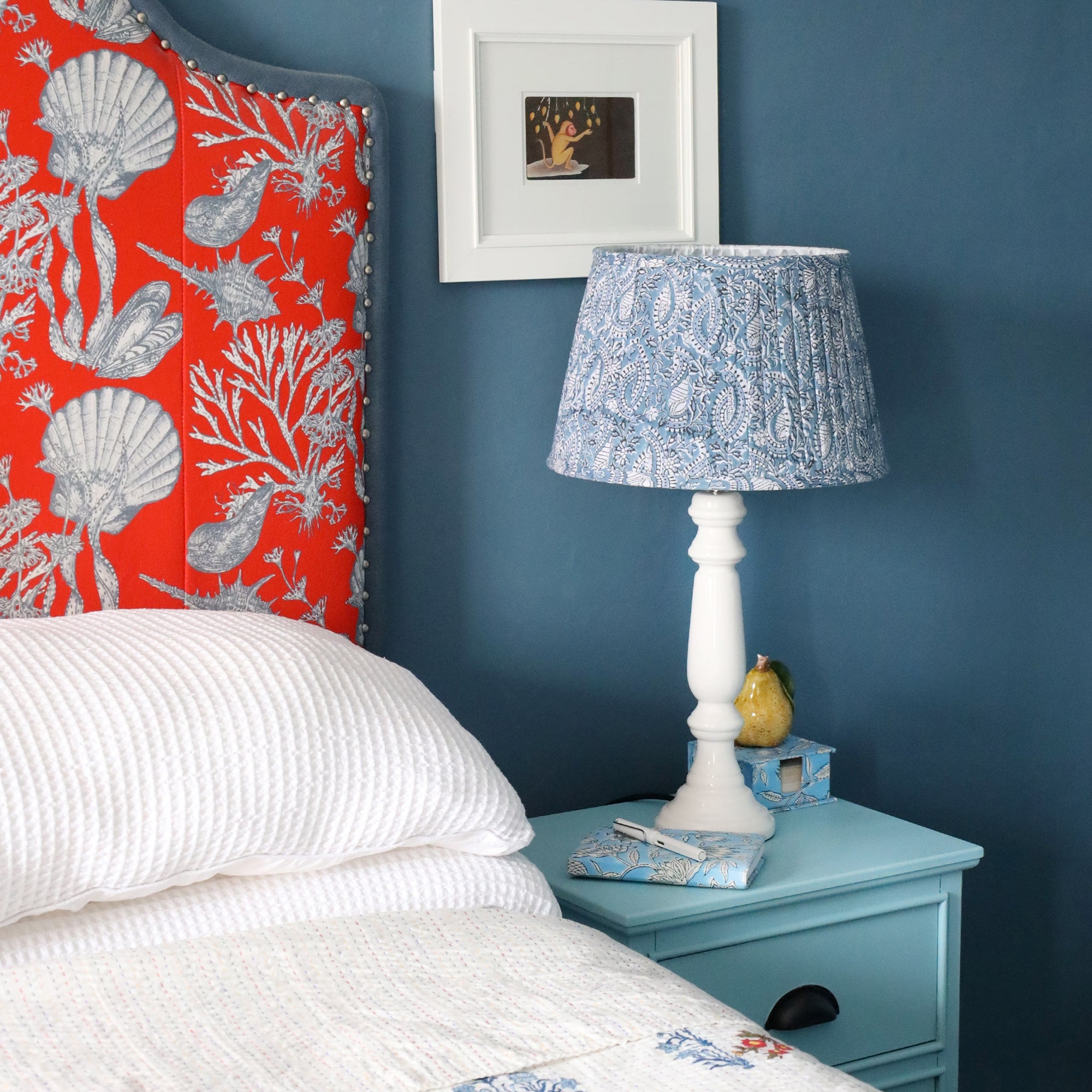 Azure Paisley Shell pleated lampshade with a white lampbase,on a bedside cabinet with a notebook and pen.In the background is the headboard which is red with white shells and a white picture on the wall.