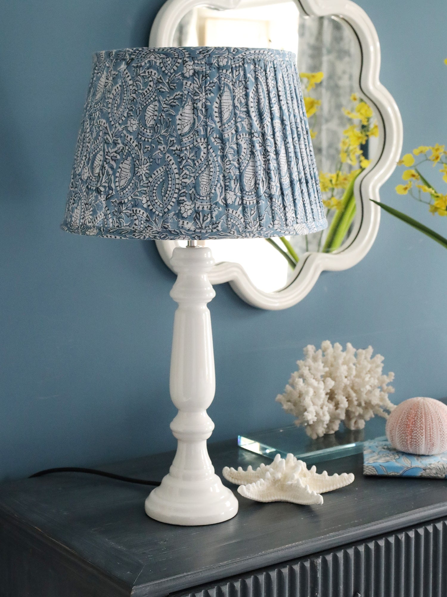 Azure paisley pleated lampshade on a wood white zennor lampbase on a sideboard.Also on the sideboard are many shells and in the background on the wall is a white wave mirror
