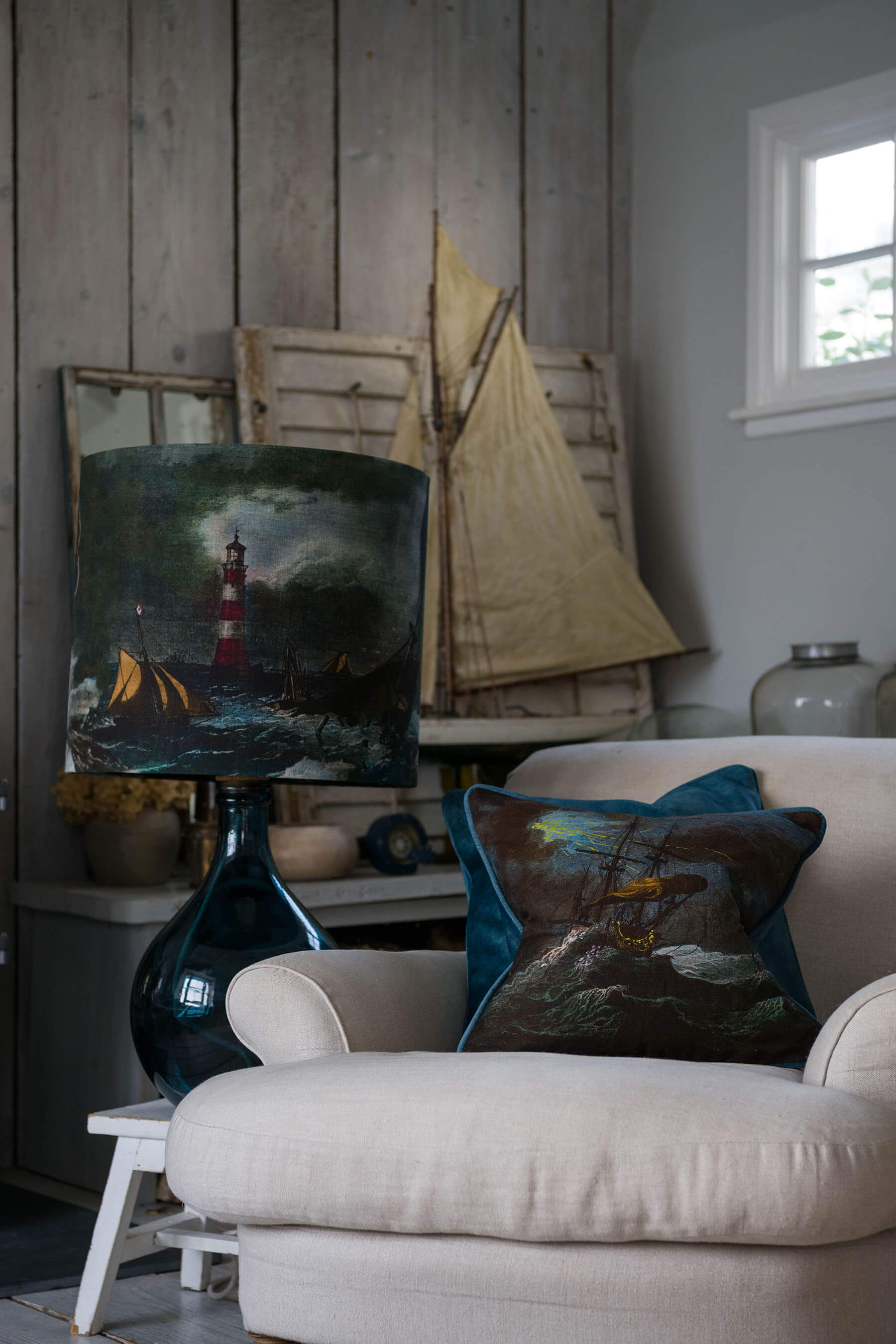 Shipwreck night cushion placed on a cream chair in front of a decorative sailing boat and other decorative items on a side table.Next to the chair is a matching lampshade on a petrol glass lampbase.