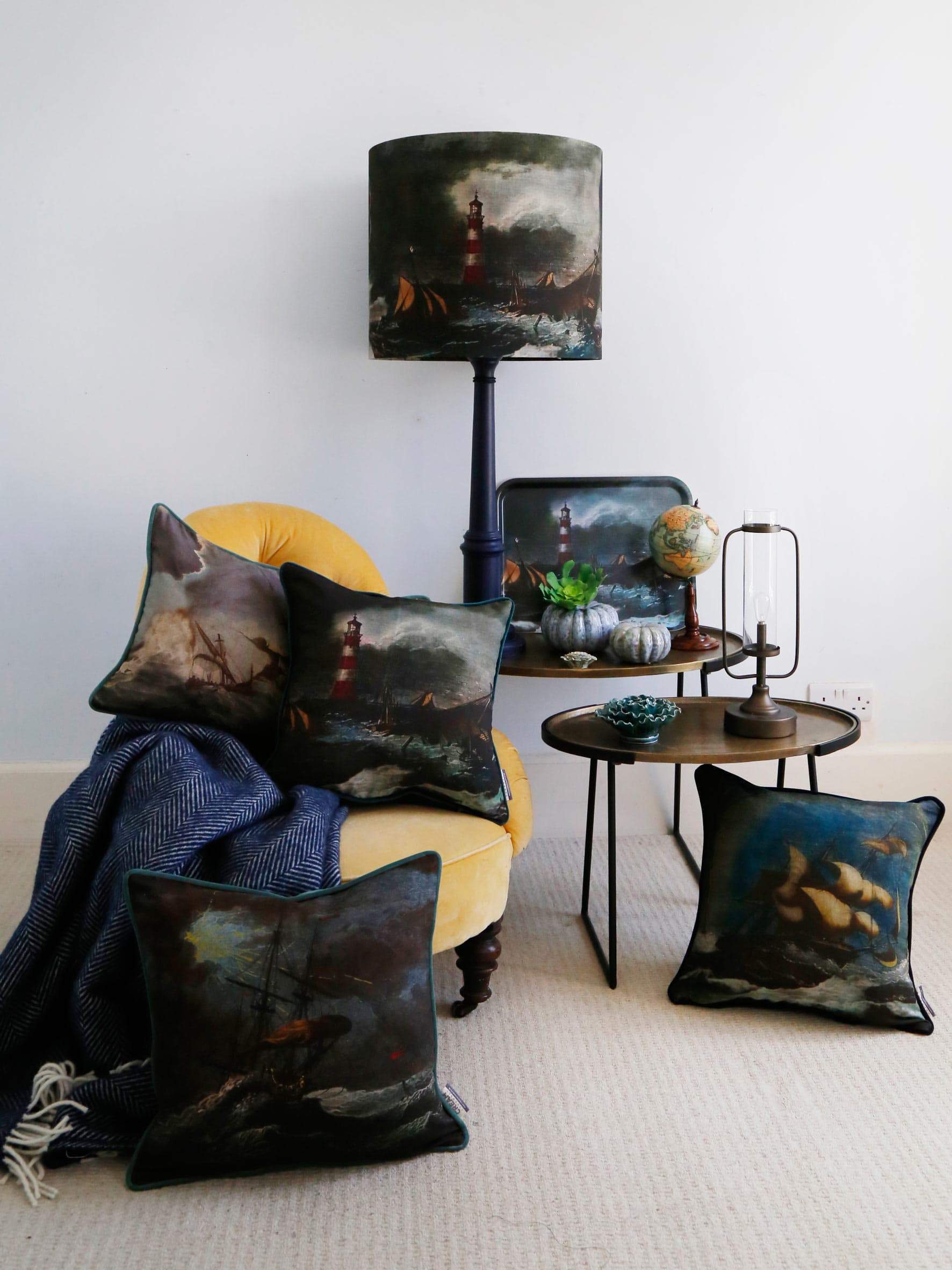 Shipwreck Night Cushion Square Cover placed on a chair with other shipwreck cushions.All piled with a navy throw all placed on a mustard velvet chair.Behind the chair is a table with a tall navy lampbase and other decorative items