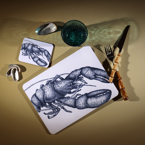 Lobster Design in Navy on a white Coaster with a matching Placemat.On the table is a bamboo cutlery set and a blue coloured glass