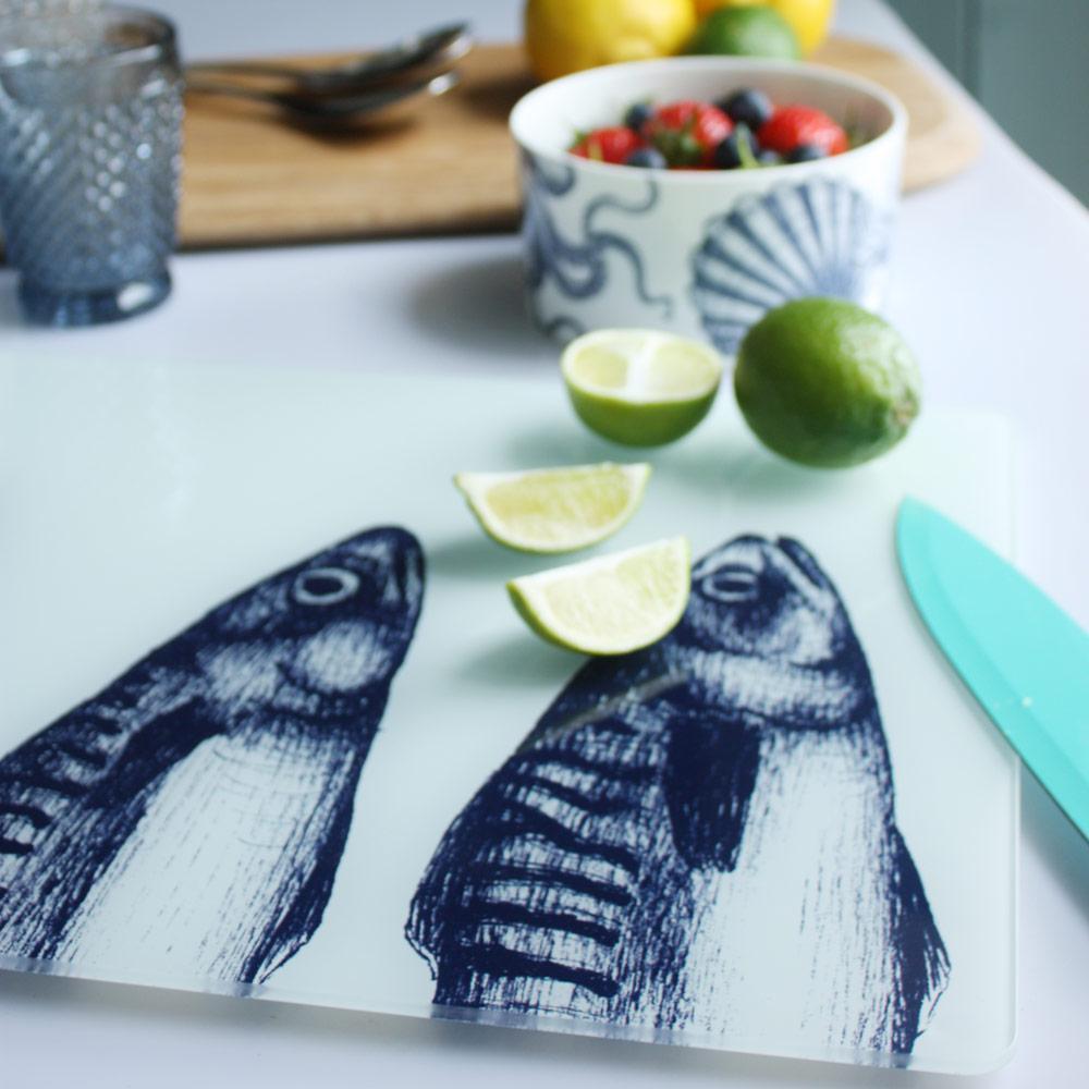 Close up of Worktop saver on toughened recycled glass with our Mackerel Heads design placed on a table with coloured glasses,a wooden board,fruits and a bowl on the table