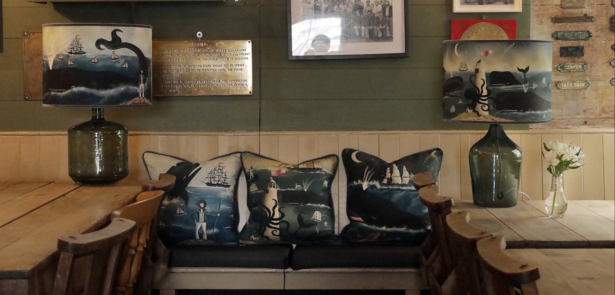 A cozy café seating area with a nautical theme, featuring two large table lamps with oceanic illustrations of whales, ships, and sea creatures. The bench is adorned with decorative cushions showcasing similar maritime designs, including ships, a lighthous