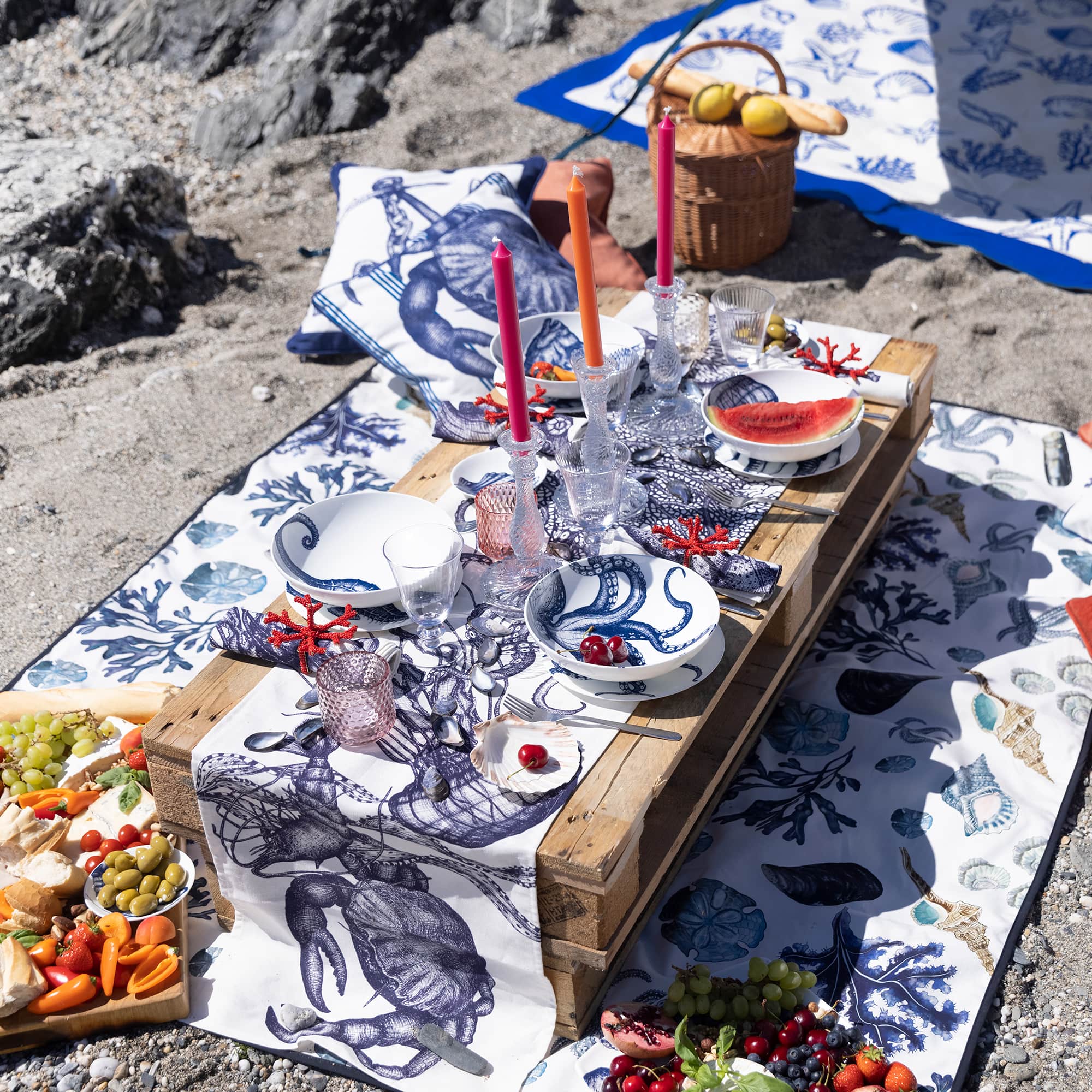 Cotton and Linen mix Sea Creature table runner displayed on a beach over a Picnic blanket.On the table runner are various Classic tableware with colourful foods in some of the bowls.In the background you can see another blanket and some sea creature cushions.On the table are various pieces of glassware and candles catching the sunlight