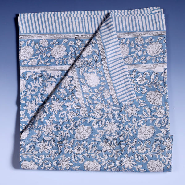 Seashell Flower tablecloth which is a  Hand block printed fabric in a soft blue and the print is swirling shells and tendrils in white on a napkin.Around the pattern is a white and blue striped finished edge.The picture shows the tablecloth folded up and a corner folded across.