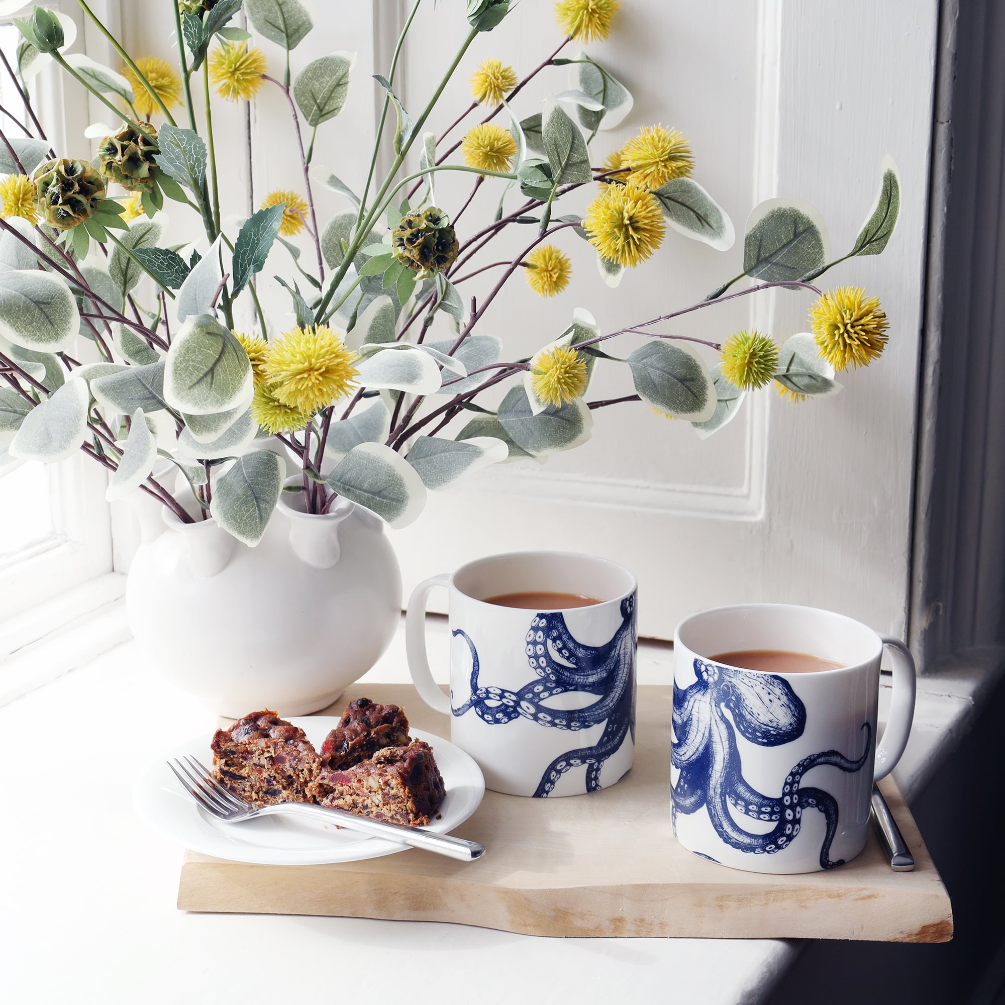 2 white bone china mugs each with a navy illustrated octopus on. They are sitting by a window on a wooden board with a vase of yellow pom pom of flowers in the background and slices of fruit cake to the side.
