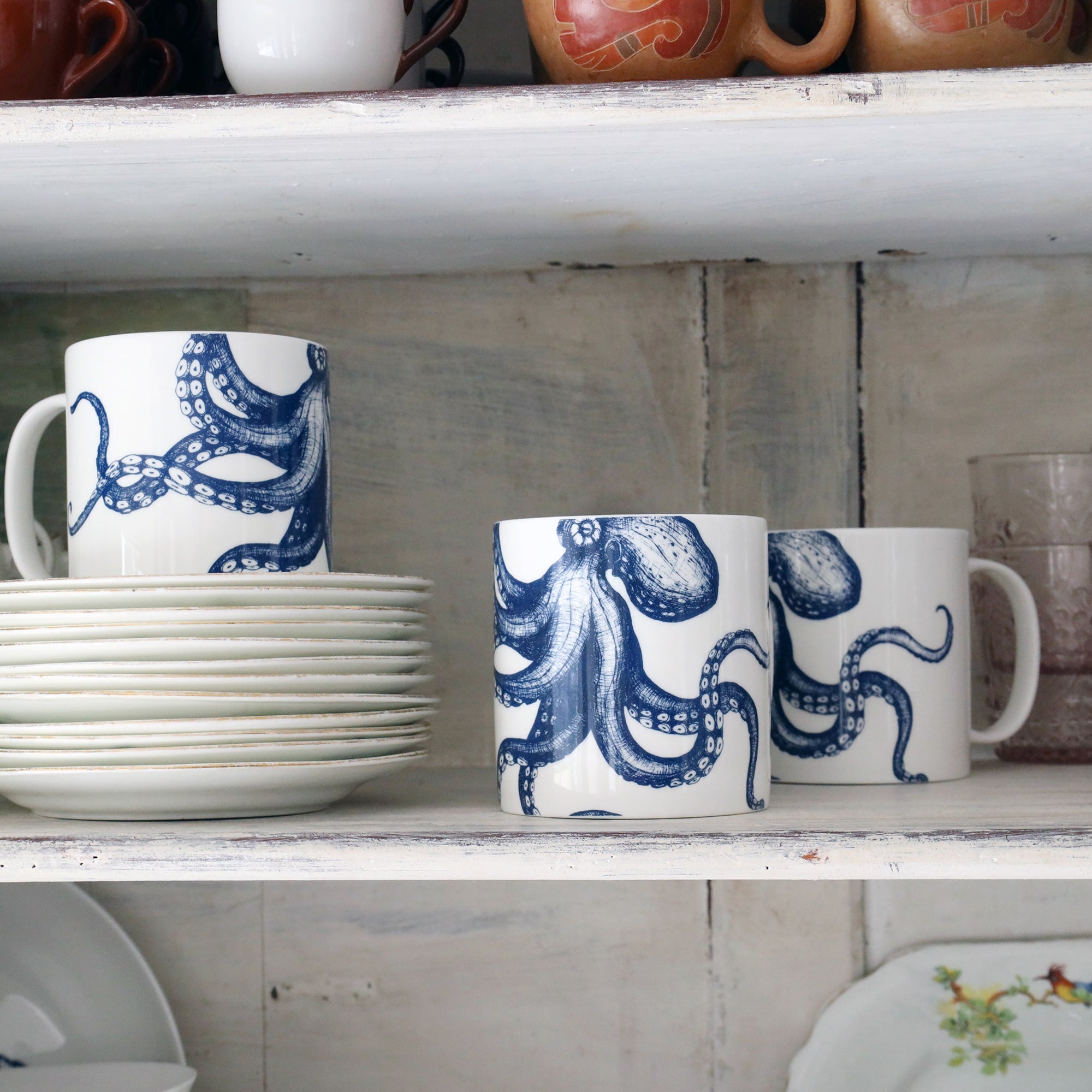 3 white bone china mugs with a navy octopus illustration on them, sitting on a cupboard shelf with other china around them.