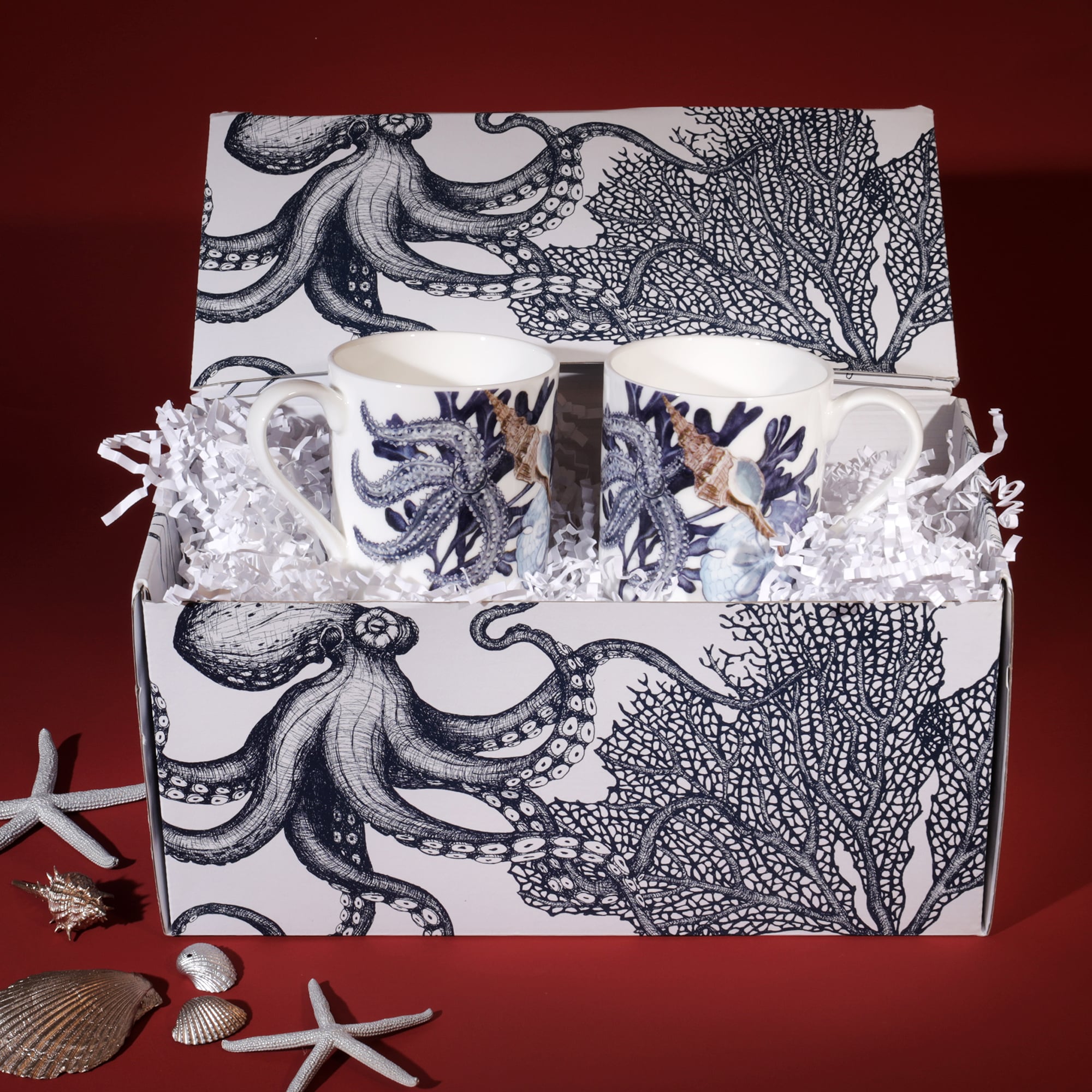 A Cream Cornwall gift set featuring two ceramic mugs with octopus and seashell designs. The mugs are presented in a matching gift box adorned with intricate illustrations of an octopus and coral. The box is filled with white shredded paper for cushioning, and decorative seashells and starfish are arranged around it, complementing the coastal theme against a deep red background.