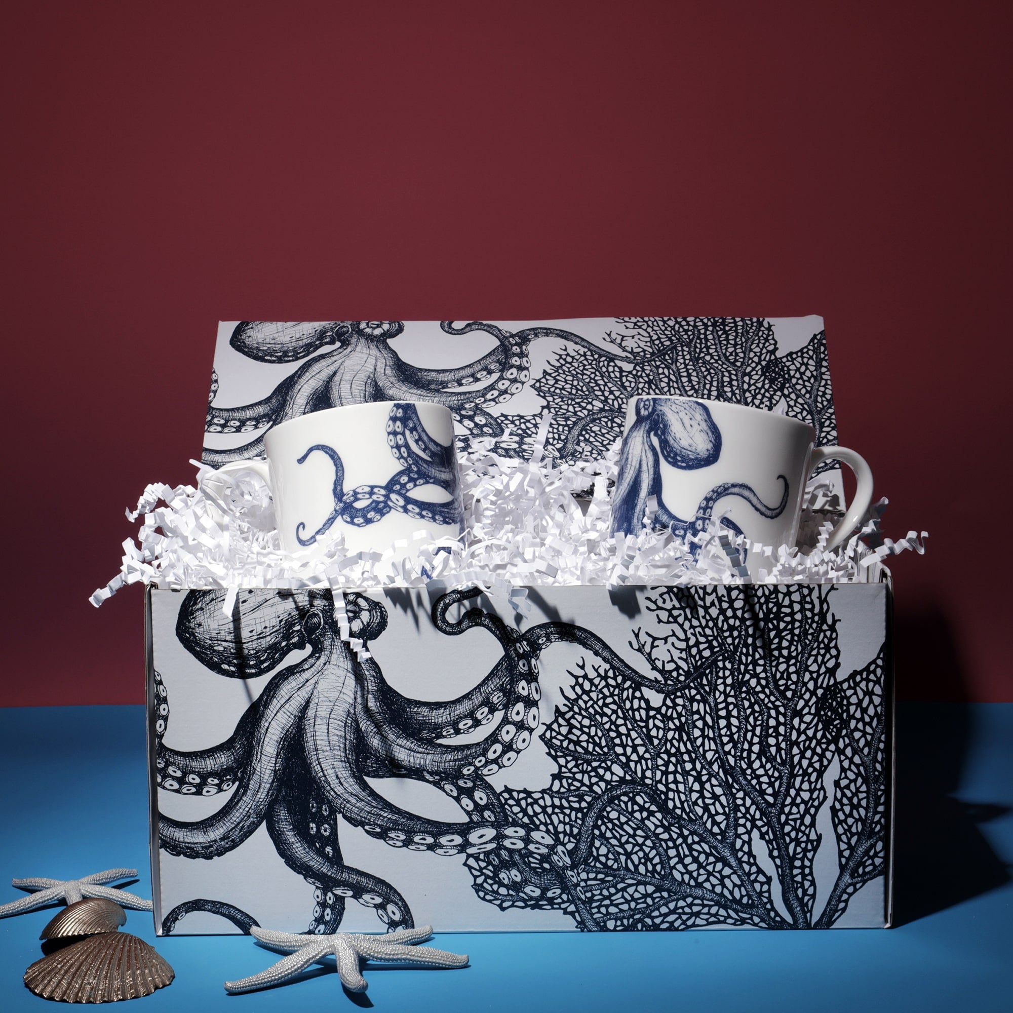 A Cream Cornwall gift set featuring two ceramic mugs with blue octopus designs. The mugs are presented in a matching gift box adorned with intricate octopus and coral illustrations. The box is filled with white shredded paper for cushioning, and decorative seashells and starfish are arranged around it, enhancing the coastal theme against a deep red and blue background