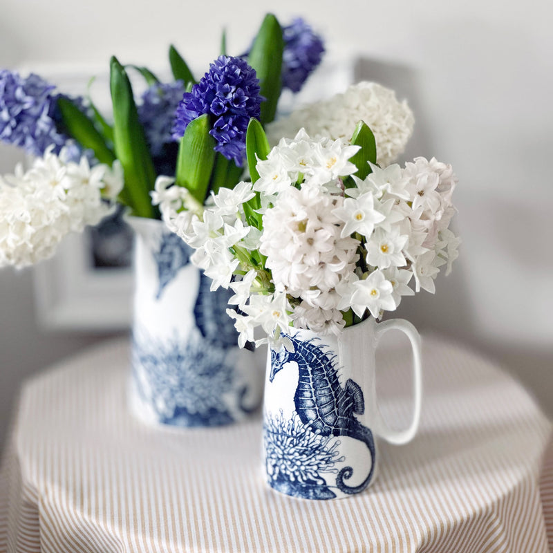 2 white bone china jugs with seahorse and anemone navy illustrations decorating them. They have hyacinths and small white spring daffodils in them and are sitting on a natural coloured striped fabric.