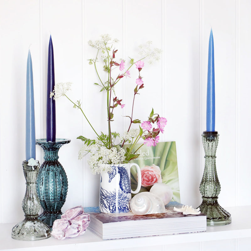 3 blue toned taper dinner candles win glass candle sticks, sitting on a white shelf with books and a blue and white seahorse jug with wild flowers