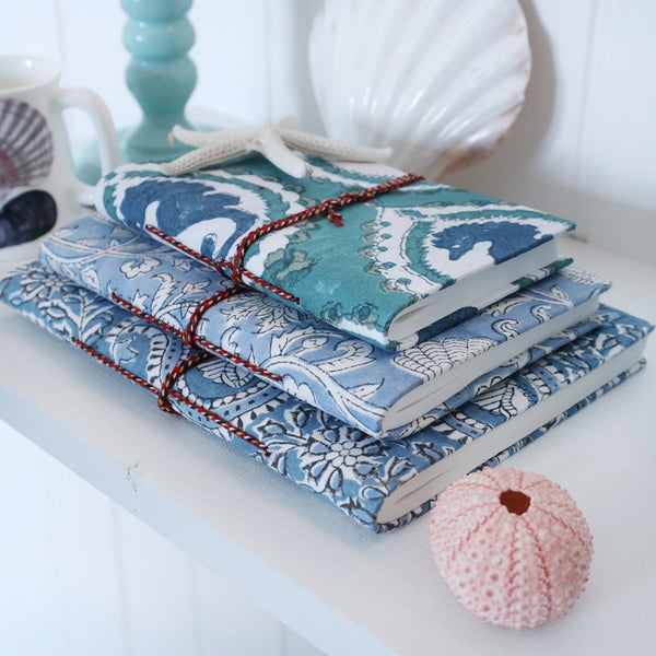 Hand Block printed hard backed notebooks stacked on a shelf.There are shells,a candlestick and a beachcomber jug also on the shelf.