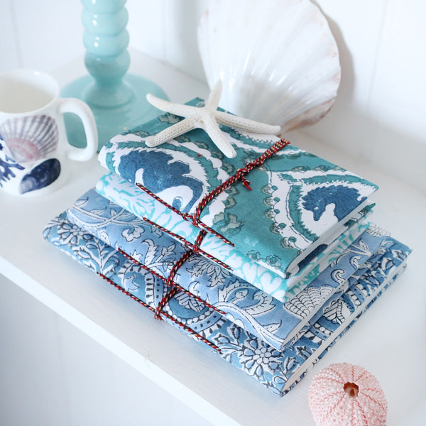 Hand Block printed hard backed notebooks stacked on a shelf.There are shells,a candlestick and a beachcomber jug also on the shelf.