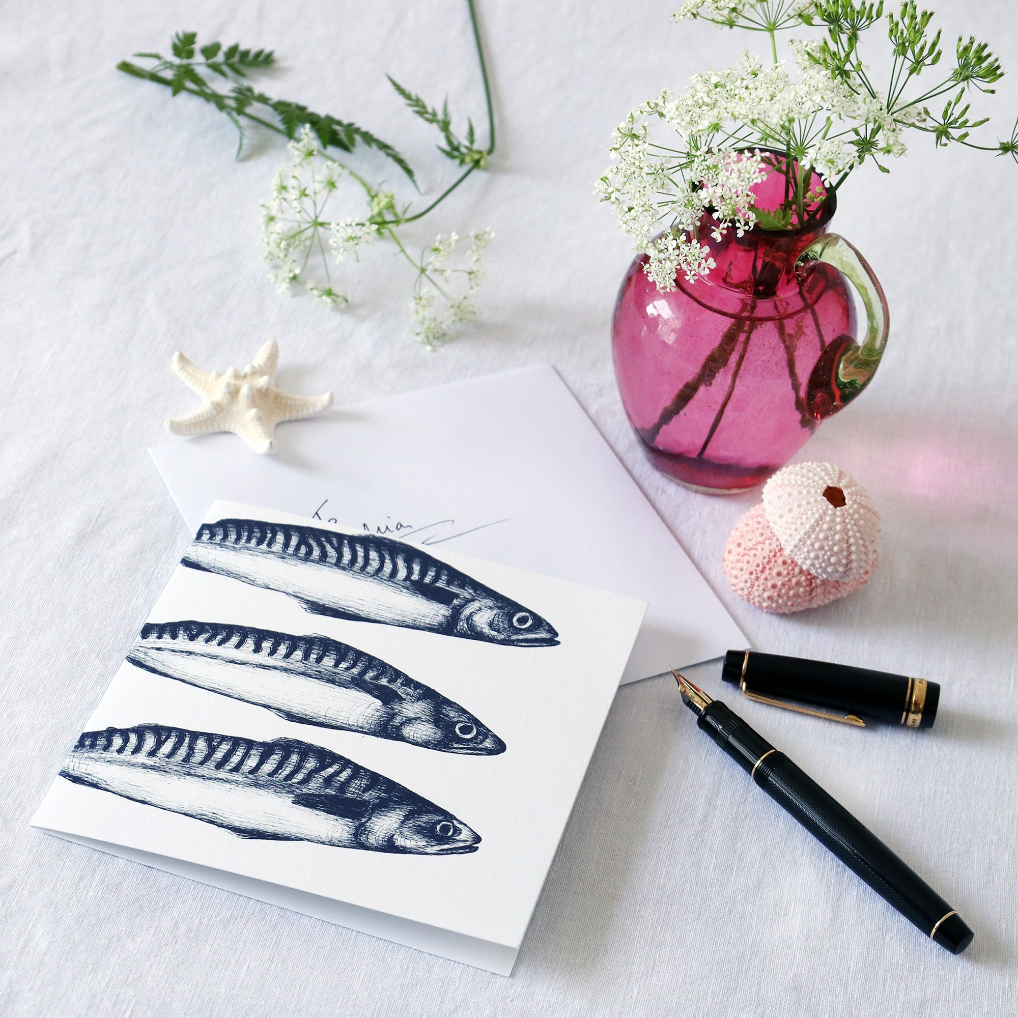 greeting card with navy illustration of 3 mackerel without tails on a white background lying on a white table cloth with a fountain pen, hand written envelope shells and a small cranberry glass jug with wild flowers in