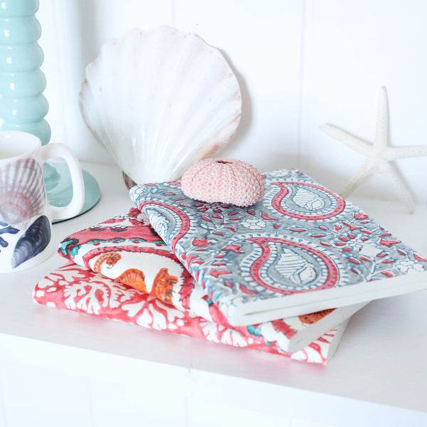 Hand Block printed paper backed notebooks stacked on a shelf.There are shells,a candlestick and a beachcomber jug also on the shel