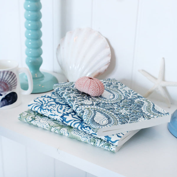 Hand Block printed paper backed notebooks stacked on a shelf.There are shells,a candlestick and a beachcomber jug also on the shelf.
