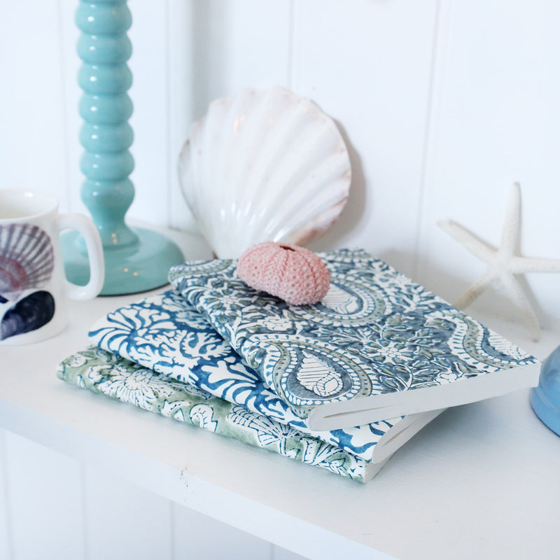 Hand Block printed paper backed notebooks stacked on a shelf.There are shells,a candlestick and a beachcomber jug also on the shelf.