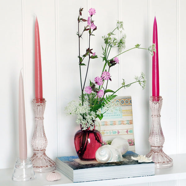 3 pink toned taper dinner candles in glass candle sticks, sitting on a white shelf with books and a cranberry glass jug with wild flowers