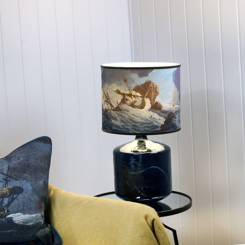 A shipwreck scene lampshade in blues & brown sitting on a petrol blue glass lamp base, set against a white shiplap wall a mustard throw over the chair and the corner of a matching cushion.