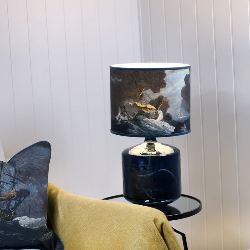 A shipwreck scene lampshade in dark blues & brown sitting on a petrol blue glass lamp base, set against a white shiplap wall a mustard throw over the chair and the corner of a matching cushion.