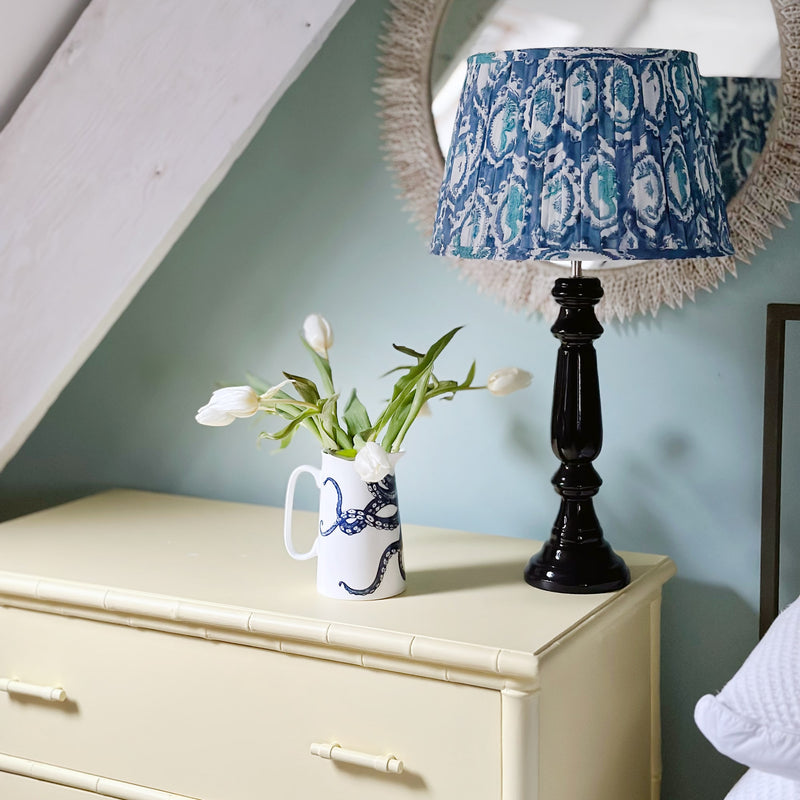 A white jug with a navy illustrated octopus on it, with a few white tulips. This is sitting on a soft yellow set of drawers and next to it is a navy lamp with a blue block printed lampshade and a mirror decorated in cut white shells.