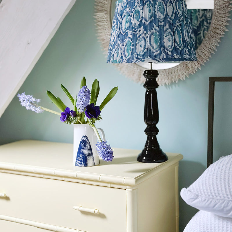 A white jug with a navy mackerel heads coming up from the bottom of the jug with blue hyacinths and anemones in it. This is sitting on some soft yellow drawers next to to a bed with a navy lacquered lamp base and blue block printed lampshade. There is a mirror behind the lampshade that is decorated in cut white shells.