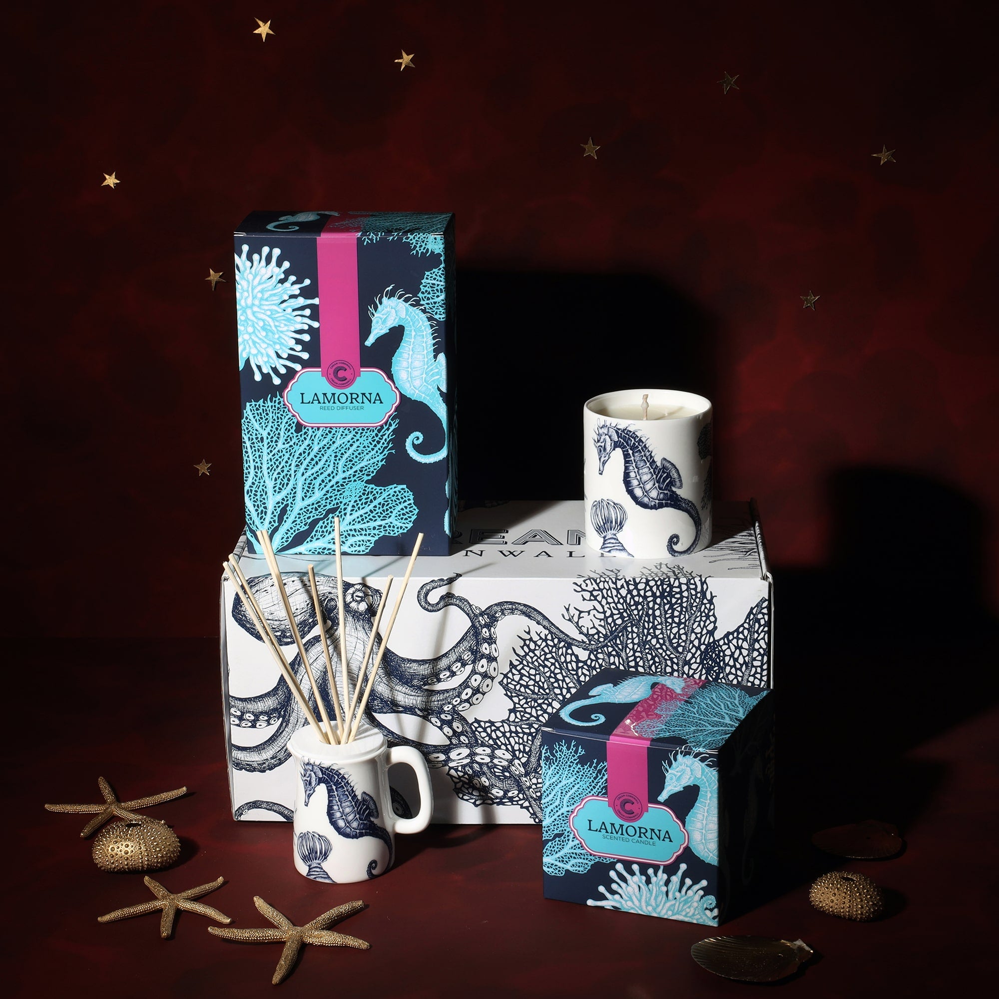 A Cream Cornwall Lamorna-themed fragrance gift set, featuring a reed diffuser and a scented candle. The packaging and candle holder are adorned with illustrations of seahorses and coral in teal and navy. The products are displayed on a box decorated with octopus and coral designs. Decorative starfish and seashells are scattered around the set, enhancing the coastal theme against a deep red background with small gold stars